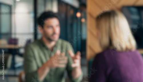 Blurred image depicting two individuals engaged in a conversation indoors. One person gestures while the other listens attentively in a modern office environment.