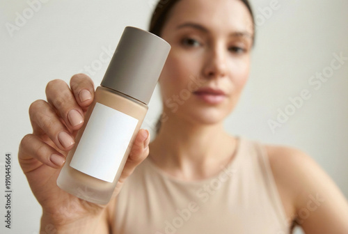Close up of hand holding foundation bottle with blank label against blurred face of beautiful woman