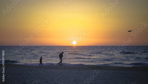 A father and child walk along the beach at sunset against the background of a calm horizon
