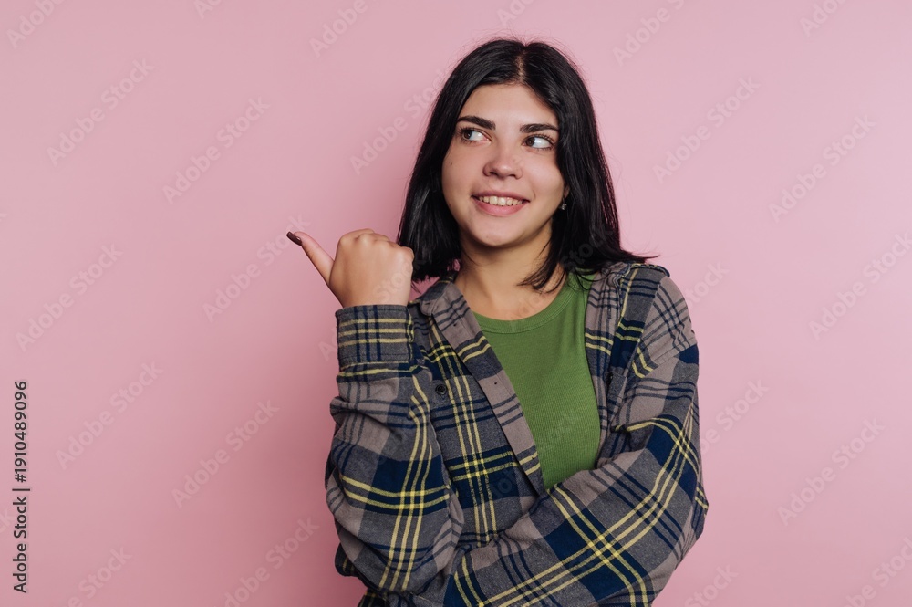 Fototapeta premium Smiling young woman in a casual flannel shirt gestures playfully against a soft pink background