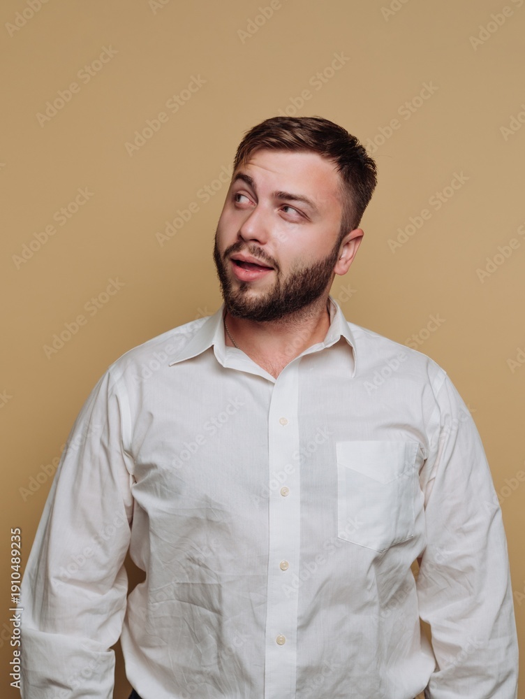 Obraz premium Young man with a beard in a white shirt gazes thoughtfully against a warm beige background