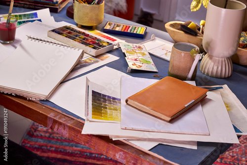 Creative artist workspace with watercolor palette, sketchbook, color swatches and notebook on messy table in cozy studio. Concept of creativity, inspiration, art education and handmade design process