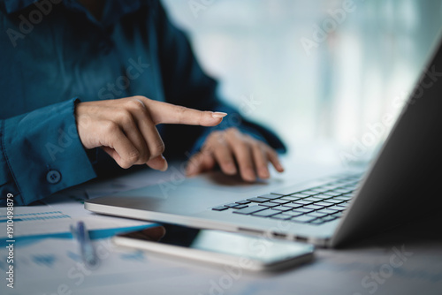A woman is pointing at a laptop screen while sitting at a desk with a cell phone