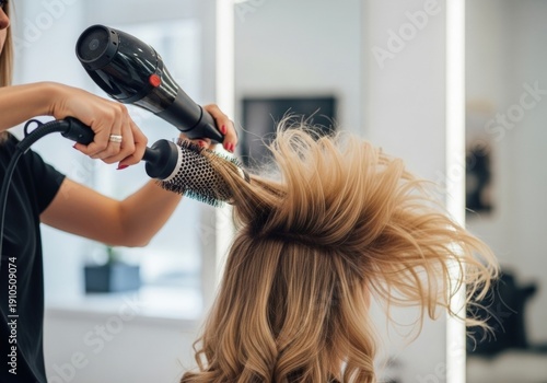 Dynamic action shot of a stylist using a round brush and a professional hairdryer to create a bouncy blowout on a woman with long blonde hair.