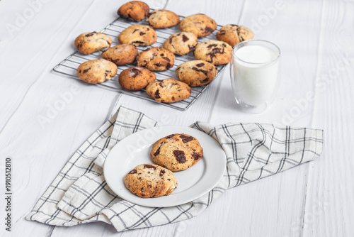 Fresh homemade chocolate chip cookies on a plate with milk and cooling rack on light wooden background