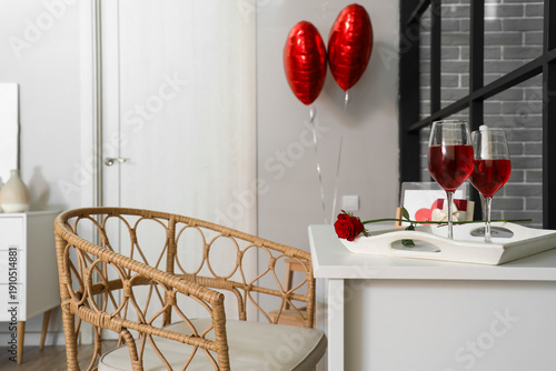 Tray with red rose and glasses of wine on table in room decorated for Valentine's Day
