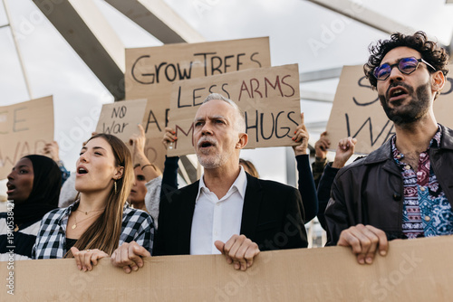 Demonstrators of varying ethnicities and ages passionately protesting for peace and anti-war causes, holding cardboard signs with messages like no war and grow trees less arms more hugs