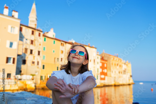 Funny little girl posing on embankment sunny summer day. Rovinj town in background. Travel and adventure concept.
