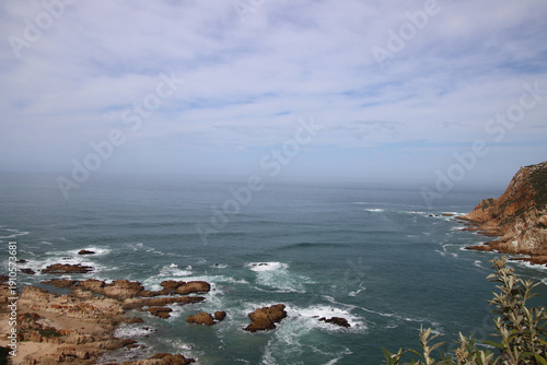 Wide view over the Indian Ocean with rocky shoreline and a coastal headland near Knysna, South Africa, under a hazy, partly cloudy sky.
