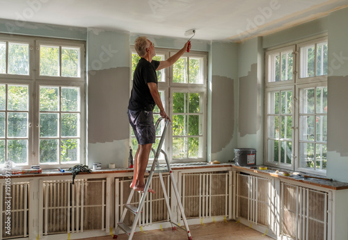 Retired man painting ceiling while standing on ladder at home