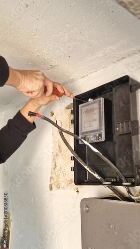 Vertical, close-up of worker hands using a screwdriver to connect electrical wires to a new digital meter box. The installation is on a white concrete wall during a home renovation project.