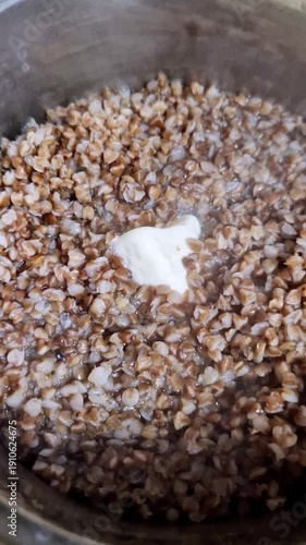 Vertical, extreme close-up overhead shot of a dollop of white butter slowly melting into a pot of hot, steaming, cooked buckwheat grains. The process creates a savory and rich texture.