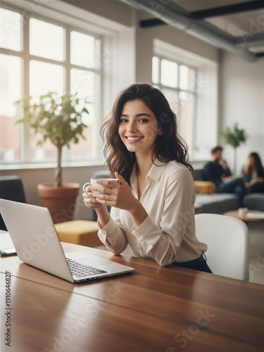 Smiling female employee sitting at wooden desk with laptop in modern open plan office, holding coffee cup, break time, professional woman, career satisfaction, workplace wellness, candid portrait.