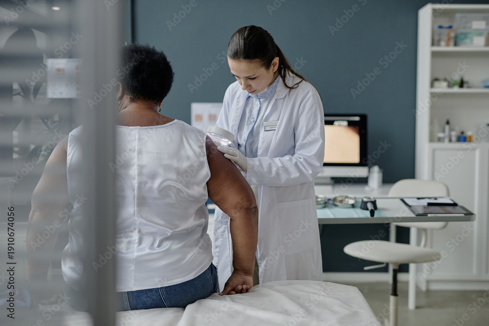 Obraz premium Senior Black woman sitting on examination table while young adult Caucasian female dermatologist examining skin on upper arm using dermatoscope in medical clinic