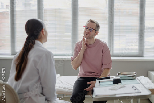 Caucasian middle aged man touching neck while consulting female doctor in medical office, discussing dermatological symptoms during examination, clinical setting visible in background