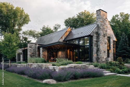 Modern stone house located in a green area with large windows and a stone chimney and surrounded by grass and plants in bright daylight