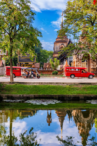 Traditional Lanna style pagoda of Wat Lok Moli temple reflected in moat water with iconic red songthaew trucks and tuk-tuk driving past. Famous landmark in Chiang Mai, Thailand at daytime