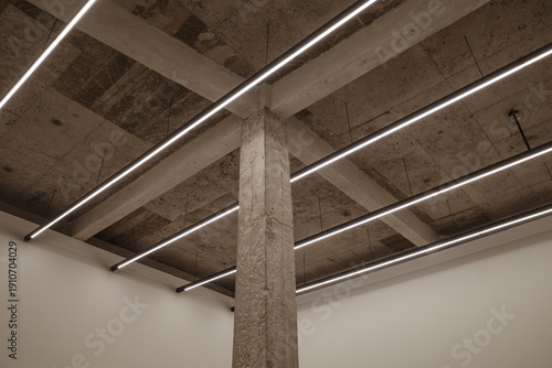 A modern interior shot featuring a raw concrete pillar and ceiling with exposed structural beams. Bright linear LED light fixtures are suspended in a parallel arrangement.