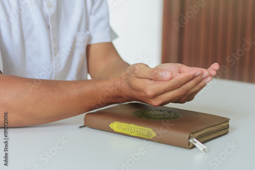 Muslim man holding hands in prayer over closed Quran on white table. Concept of faith, peace, devotion, spiritual growth, and Islamic education during the holy month of Ramadan