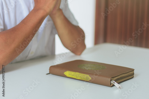 Muslim man holding hands in prayer over closed Quran on white table. Concept of faith, peace, devotion, spiritual growth, and Islamic education during the holy month of Ramadan