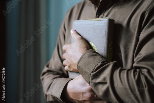 Muslim man in brown shirt holding and hugging Quran book to his chest. Concept of faith, peace, devotion, spiritual growth, and Islamic education during the holy month of Ramadan