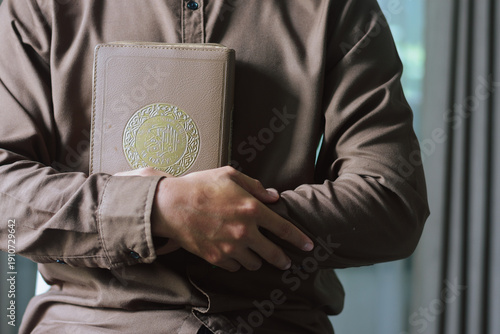Muslim man in brown shirt holding and hugging Quran book to his chest. Concept of faith, peace, devotion, spiritual growth, and Islamic education during the holy month of Ramadan
