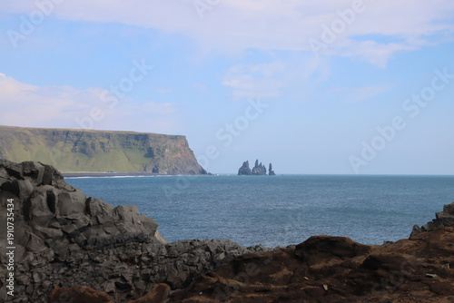 View from Dyrholaey over the Atlantic Ocean toward Reynisdrangar basalt sea stacks near Reynisfjara on Iceland’s south coast, with dark volcanic rocks in the foreground.