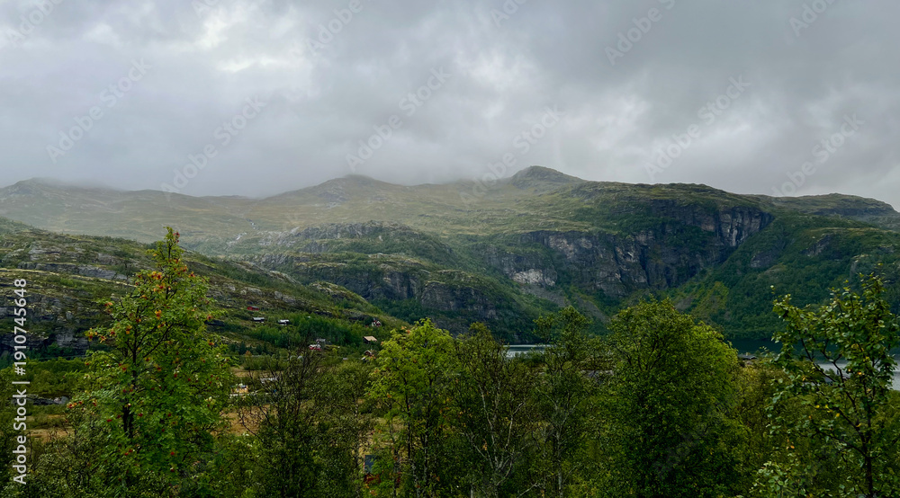 Fototapeta premium Dramatic mountain landscape in Myrdal, Norway featuring steep rocky cliffs, lush green valley, scattered cabins, and misty clouds rolling over the peaks, showcasing the raw beauty of Scandinavian natu