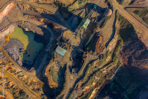 Aerial view of earthworks scarred with raw umber gashes, a metallic structure standing stark against ochre tones, and pools of jade water, La Roche-Jaudy, Brittany, France.