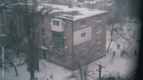 Snow falls on a Soviet-era apartment block as people and a car move below. Static high-angle shot with a desaturated look captures a bleak, cold urban atmosphere.