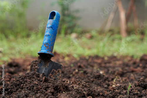 A low-angle, close-up shot of a small black and blue plastic hand trowel covered in moist, dark potting soil, embedded in a newly tilled garden bed. The background features soft focus greenery, emphas