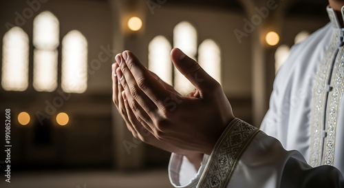 A person's hands in a prayer gesture, palms open, wearing a white robe with ornate sleeves, inside a dimly lit religious building with arched windows.