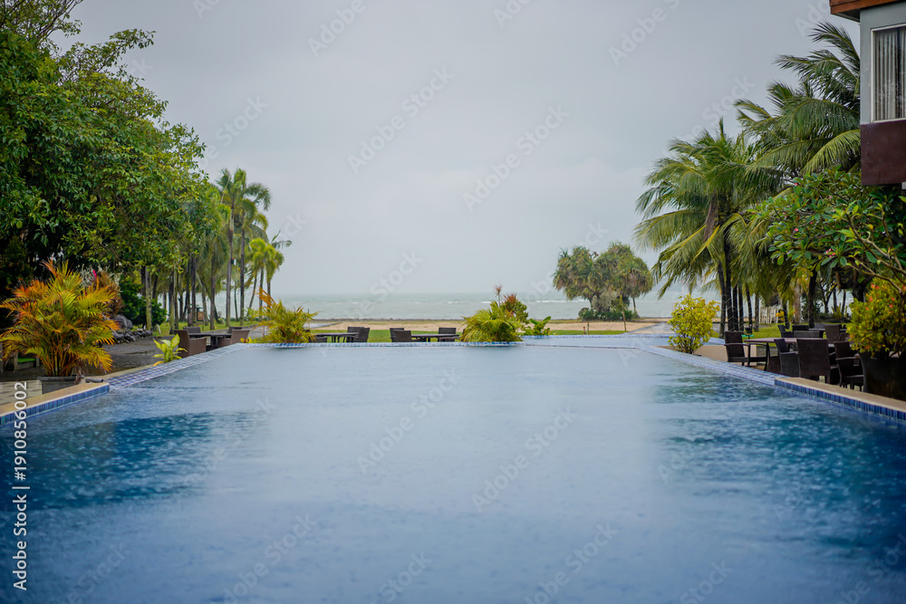 Obraz premium Infinity pool facing the beach on a rainy day, surrounded by tropical trees and lush greenery