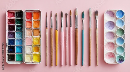 Colorful Watercolor Tools and Palette Arranged Horizontally on a Soft Pink Background for Artistic and Creative Imagery