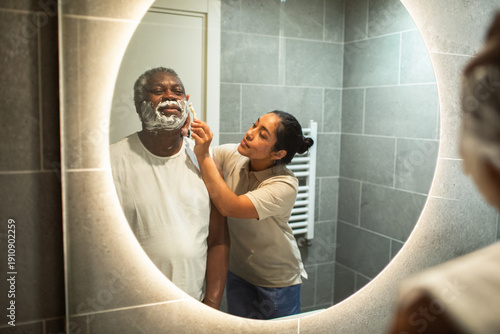 Caregiver helping senior man shave in bathroom mirror