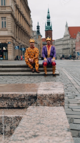 Two men in fancy dress and crown costume sitting on stone steps in an old European city square. Three kings concept for Epiphany.