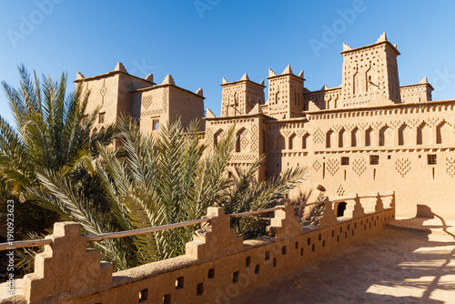 Exterior view of the Kasbah Amridil, a traditional earthen building with towers and geometric designs, framed by palm trees against a clear blue sky. Kasbah Amridil,Marrakech,Morocco