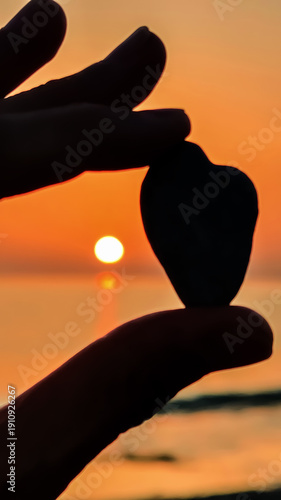 Holding a heart-shaped stone against the backdrop of a sunset over the water creating a moment of connection and reflection