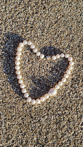 Pearl necklace arranged to form a heart shape on sandy beach during sunny day with natural light casting shadows