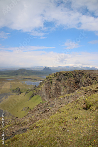 Wide elevated view of rugged cliffs and green volcanic highlands near Dyrholaey on the south coast of Iceland, overlooking vast plains and distant glaciers under a partly cloudy sky.