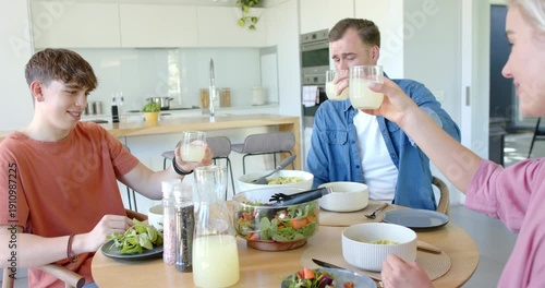Family of three raising short glasses for toast, clinking and sipping juice at modern kitchen table