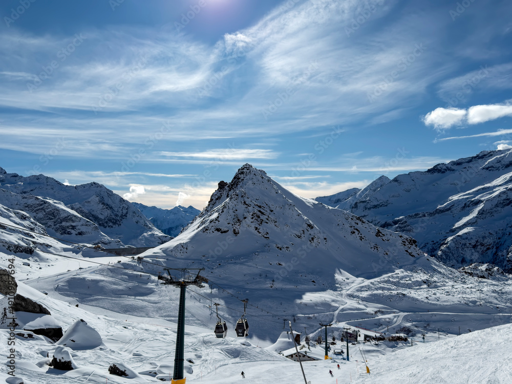 Fototapeta premium Whispers of Winter in the High Alps, A vast snowy slope stretches toward rugged alpine peaks, partially veiled by drifting clouds under a deep blue sky. The scene captures the serene beauty and raw ma