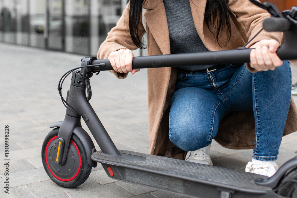 Obraz premium Young woman in a beige coat folds an electric scooter in the middle of a city street. A person uses an environmentally friendly form of transport in the form of an electric scooter.