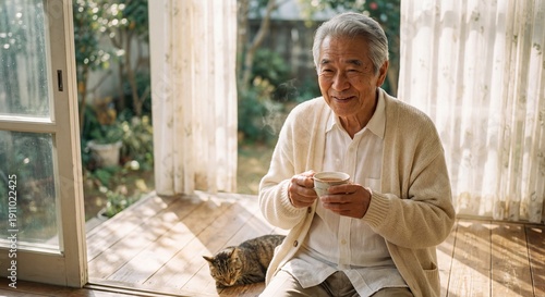 Smiling senior Asian man holding warm tea cup on sunny veranda with cat resting at his feet and garden in background