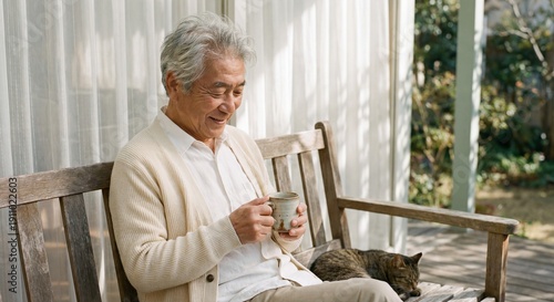 Senior Asian man smiling while holding ceramic tea cup on wooden porch bench with sleeping tabby cat