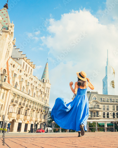 Young woman tourist in flowing blue dress and summer hat running across sightseeing Europe Square in Batumi, Georgia. Vibrant tourism and culture at the Black Sea resort city Batumi