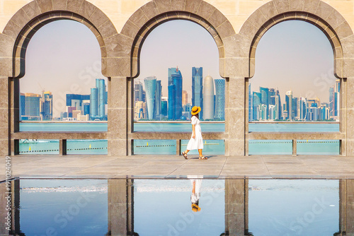 Woman tourist wear dress walking near Museum Islamic Art Doha with stone arches framing modern skyline reflected in water Middle East travel lifestyle architecture landmark