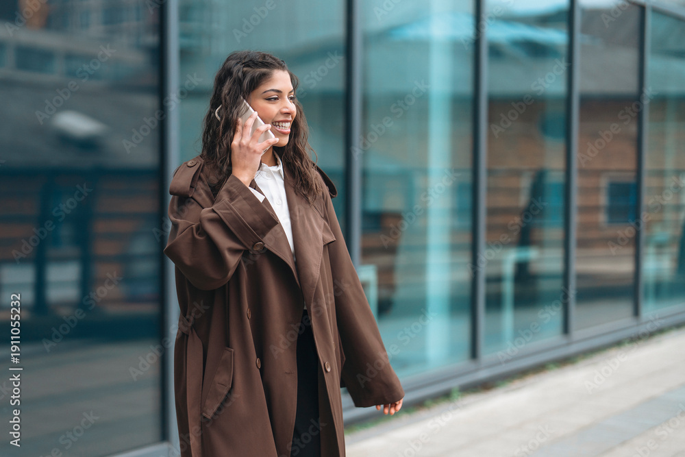 Fototapeta premium Woman talking on phone while walking near building with glass windows in city