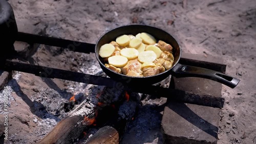 Chicken and potatoes sizzle in a pan over a crackling campfire. A rustic outdoor cooking scene. Side view.