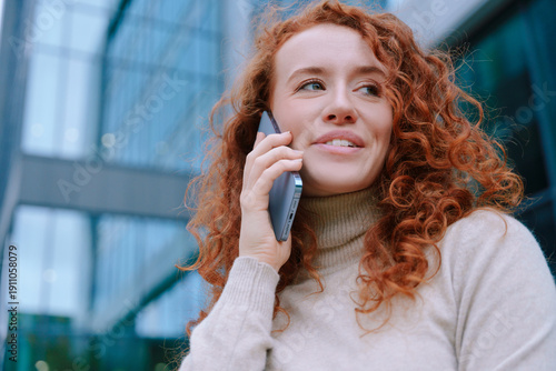 Young woman talks on her phone outside in a city during the day near glass buildings